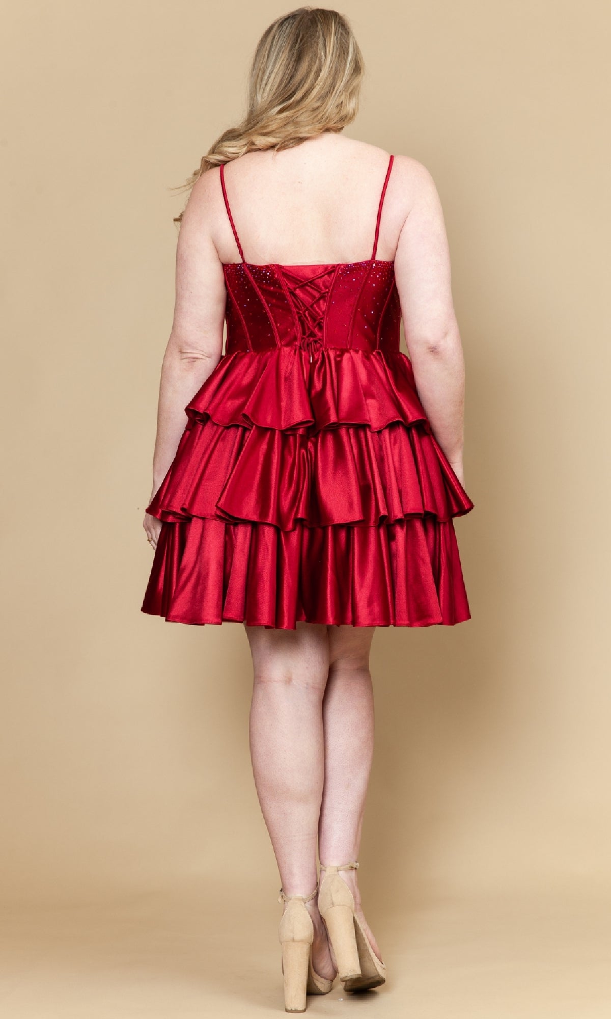 A woman with blonde hair wears the Poly USA W1192 Short Plus Size Homecoming Dress, a sleeveless tiered red satin dress, and beige heels, standing with her back to the camera against a beige background.